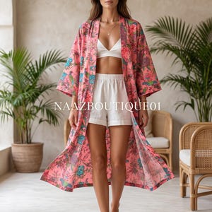 May include: A long, open-front kimono robe in a vibrant pink fabric featuring a multi-coloured floral and bird print. The garment is worn over a white bralette and white shorts. The text NAAZBOUTIQUE01 is visible across the centre of the robe.
