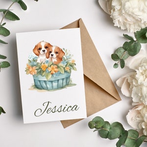 May include: A greeting card with a watercolor illustration of two brown and white puppies in a blue basket with yellow flowers. The card is personalized with the name "Jessica".