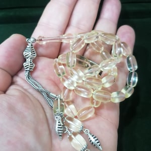 May include: A clear amber prayer beads necklace with silver accents. The beads are oval shaped and the necklace has a tassel with a silver charm.
