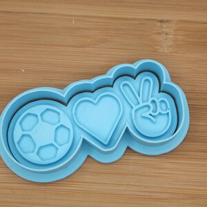 Può includere: Un tagliapasta in plastica azzurra con un pallone da calcio, un cuore e un simbolo di pace. Il tagliapasta è realizzato in plastica ed è progettato per ritagliare forme da pasta frolla o fondente. Il design combina sport e pace.