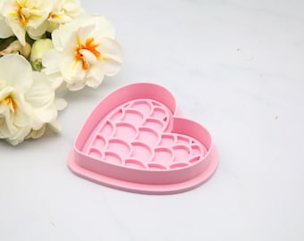 Puede incluir: Un cortador de galletas rosa en forma de corazón con un patrón interior decorativo. El cortador está sobre una superficie blanca junto a un grupo de narcisos blancos y amarillos.