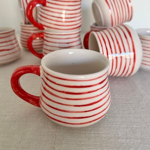 May include: White ceramic mugs with red horizontal stripes and red handles. Several mugs are stacked, and others are scattered around. The mugs are small and appear to be handmade. The mugs are on a light-colored surface.