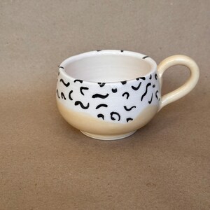May include: A white ceramic mug with a black abstract pattern and a yellow glaze on the bottom. The mug has a rounded shape and a handle.