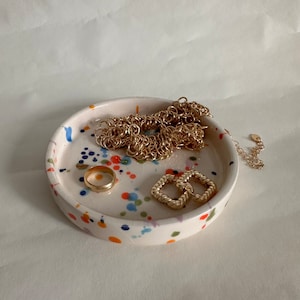 May include: A white ceramic trinket dish with a colourful splatter paint design. The dish is filled with gold jewellery, including a chain necklace, a ring, and two square earrings.