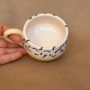 May include: A small, white ceramic mug with a yellow rim and black abstract design. The mug has a rounded shape and a handle.