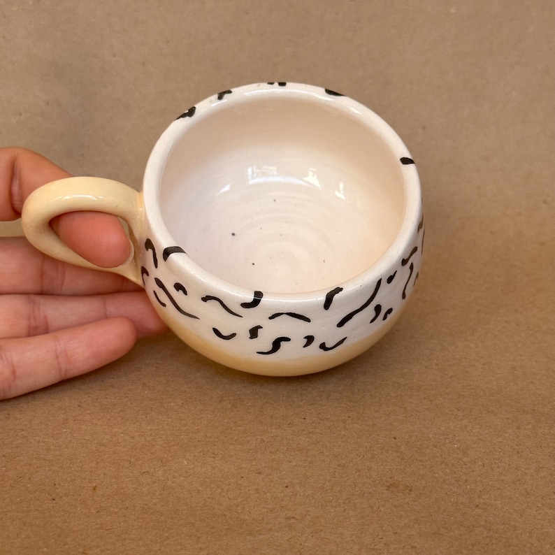 May include: A small, white ceramic mug with a yellow rim and black abstract design. The mug has a rounded shape and a handle.