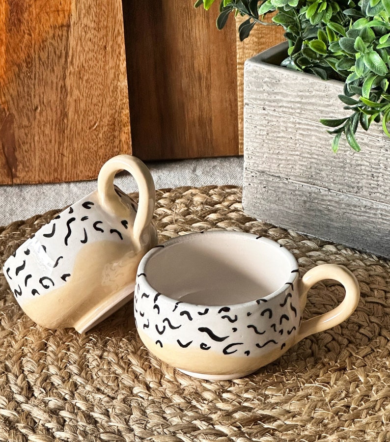 May include: Two ceramic mugs with a beige and white background and black abstract designs. The mugs have a rounded shape and a handle.