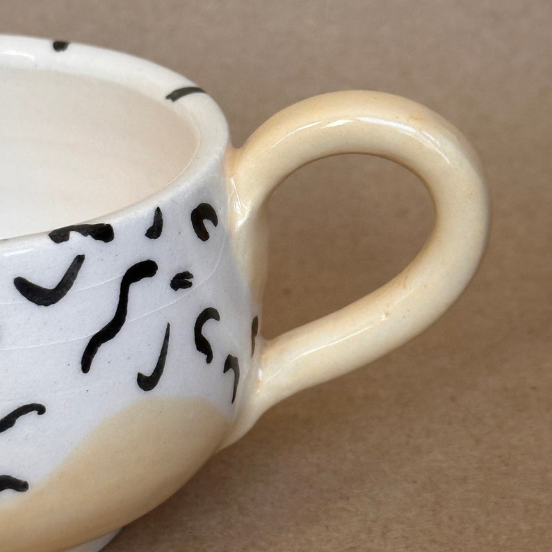 May include: A white ceramic mug with a black abstract design and a cream-colored handle.