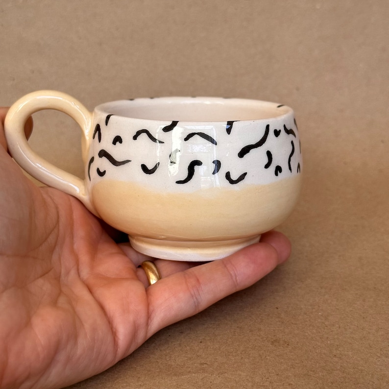 May include: A small, cream-colored ceramic mug with a black and white abstract pattern. The mug has a rounded shape and a small handle.