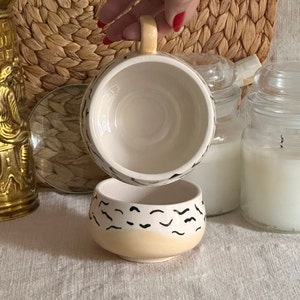 May include: A white ceramic mug with a black and yellow abstract design. The mug is upside down, revealing the inside of the mug.