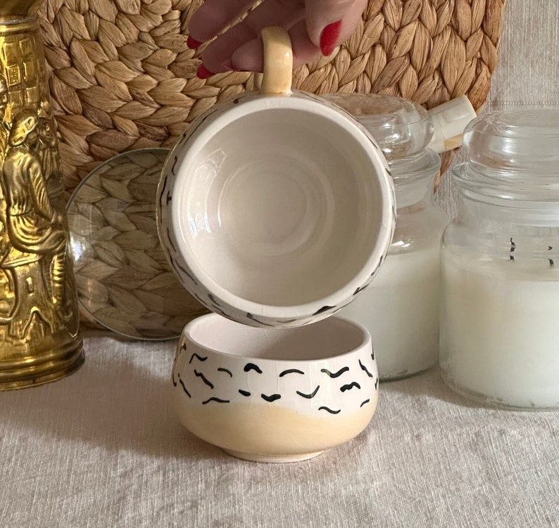 May include: A white ceramic mug with a black and yellow abstract design. The mug is upside down, revealing the inside of the mug.