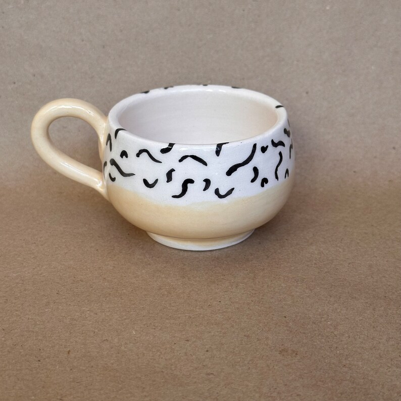 May include: A small, white ceramic mug with a black and white abstract design. The mug has a rounded shape and a handle.