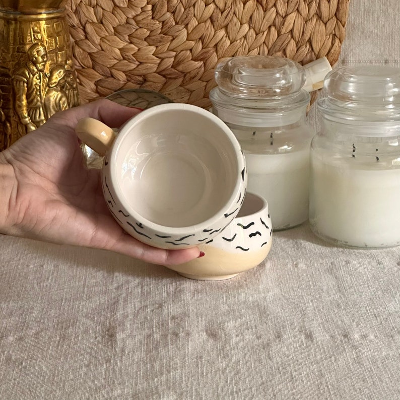 May include: A white ceramic mug with a black abstract design. The mug is partially filled with a light yellow liquid. The mug is sitting on a white surface.