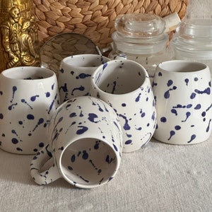 May include: Five white ceramic mugs with a blue splatter pattern. The mugs are arranged in a semi-circle on a white surface.
