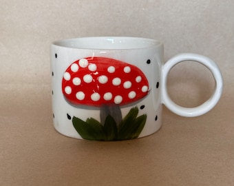 May include: White ceramic mug with a red and white mushroom design. The mushroom has white dots on a red cap and green leaves at the base. The mug has a black polka dot pattern.