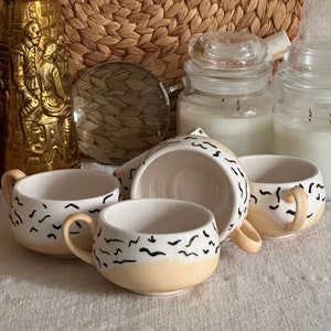 May include: Four ceramic mugs with black and white abstract designs. The mugs are white with a light beige rim and feature a wavy pattern.