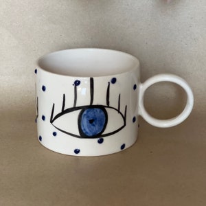 Puede incluir: Taza de cerámica blanca con un diseño de ojo azul grande y lunares azules. El ojo tiene pestañas negras y un iris azul. La taza tiene un asa redondeada, adecuada para bebidas frías o calientes.