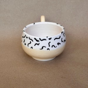 May include: A white ceramic mug with a black and white abstract design. The mug has a rounded shape and a handle.