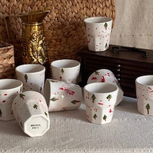 May include: Set of eight white ceramic mugs with a red and green Christmas tree design. The mugs have a textured, faceted design and are arranged on a white surface. The mugs are labeled "Mickey Arts".