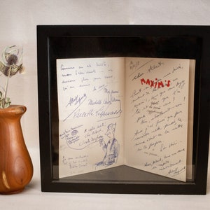 May include: A framed handwritten letter with a black frame. The letter is folded in half and has a drawing of a person on the left side. The letter is addressed to "Cher Albert" and mentions "Maxim's".