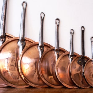 May include: A collection of copper pans with long, riveted handles. The pans are arranged in a row, decreasing in size from left to right. The copper surfaces have a shiny, reflective quality, and the handles are a darker, metallic color.