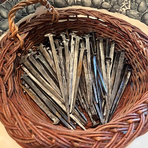 May include: A collection of antique metal nails fills a woven brown basket with a braided handle. The nails are various shades of gray and brown, with a slightly rusted appearance. The basket sits on a decorative plate with a leaf pattern.