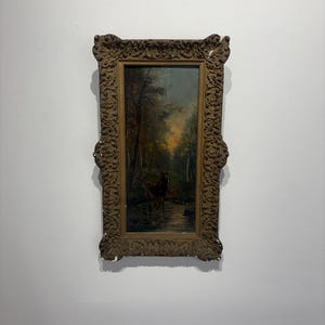 May include: A rectangular oil painting in a dark, ornate, carved wooden frame. The artwork depicts a deer standing in a shallow stream within a forest setting, featuring muted tones of brown, green, and a soft orange glow in the background sky.
