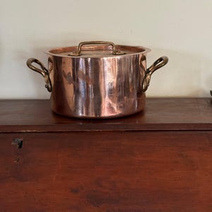 May include: A copper pot with brass handles and a lid. The pot is sitting on a wooden surface.