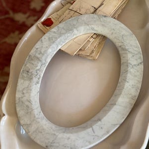 May include: Oval-shaped marble ring with gray veining. The ring is resting in a white, scalloped-edge dish. A stack of vintage letters with handwritten script and red wax seals is visible in the background.