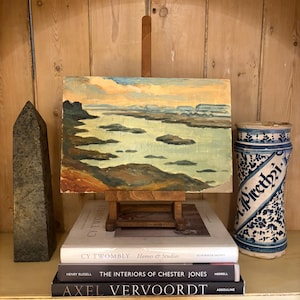 May include: An oil painting of a landscape on a small wooden easel, with a blue and yellow sky and water. A dark gray obelisk and a blue and white ceramic jar with text are on either side. Books are stacked below the painting.
