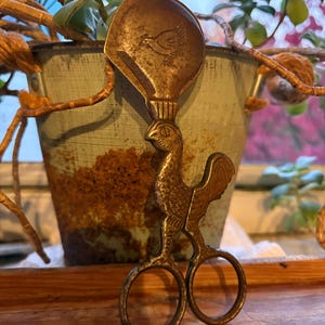 May include: Vintage metal scissor-style sugar tongs with a rooster design. The tongs have a scoop with a bird engraving and a handle shaped like a rooster. The metal has a tarnished, antique appearance, and the tongs are set against a blurred background.