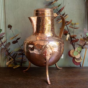 May include: A hammered copper pitcher with a lid and three legs. The pitcher has a rustic, antique look and is made of a warm, coppery metal.