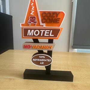 May include: A decorative sign featuring the Cozy Cone Motel logo with an orange traffic cone shape, a brown rectangular motel sign, a No Vacancy sign, and an oval sign reading 100% Refrigerated Air, all mounted on a black rectangular base.