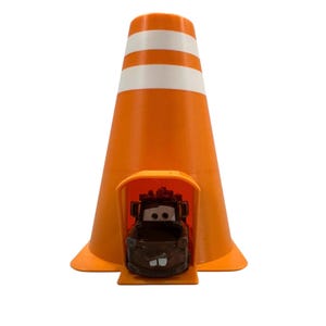 May include: A bright orange traffic cone featuring two horizontal white reflective stripes. A small brown toy truck with large eyes and a prominent front tooth is positioned inside a rectangular opening at the base of the traffic cone.
