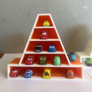 May include: A white, pyramid-shaped display shelf with orange interior shelving, holding twelve small toy cars from the Cars movie franchise. The shelf features five horizontal tiers of varying widths, organised to showcase the miniature vehicle collection.
