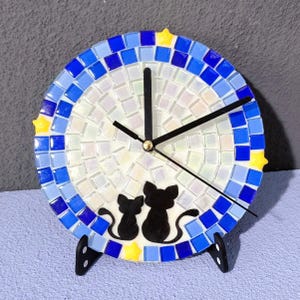 Peut inclure: Une horloge en mosaïque bleue et blanche avec des aiguilles noires et une silhouette de deux chats. L'horloge a un motif d'étoiles jaunes autour du bord.
