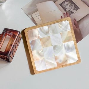 May include: A square coaster made of natural bamboo with a white mother-of-pearl inlay. The inlay is arranged in a checkered pattern.