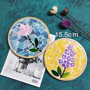 May include: Two round wooden coasters with mosaic tile designs. One coaster features a pink flower with a blue and green background. The other coaster features a purple flower with a yellow background. The coasters are 15.5 cm in diameter.