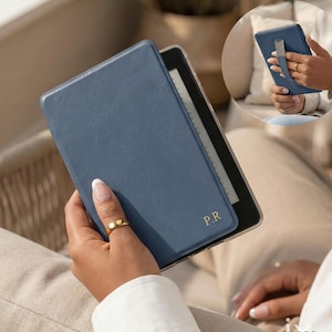 May include: A blue leather e-reader case with the initials "P.R" in gold lettering. The case is open, revealing the e-reader screen. The image shows a close-up of the case being held, with a gold ring on the person's finger.