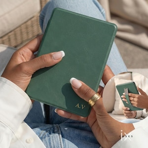 May include: A green leather e-reader case with the initials "A.V" embossed on the front. The case is rectangular and has a textured surface. The image shows the case being held by a person.