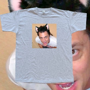 May include: A heather gray t-shirt featuring a small image of a person wearing cat ears. The person in the image is looking up at the camera. The t-shirt is plain and has a crew neck.