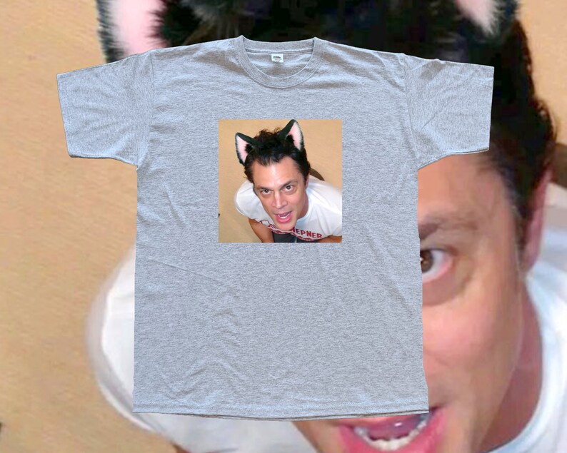 May include: A heather gray t-shirt featuring a small image of a person wearing cat ears. The person in the image is looking up at the camera. The t-shirt is plain and has a crew neck.