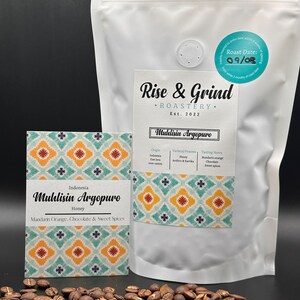May include: A white bag of coffee beans with a label that reads "Rise & Grind Roastery Est. 2022" and "Muhlisin Argopuro Honey". The label also includes details about the origin, varietal, process, and tasting notes. The bag is sitting on a black surface with coffee beans scattered around it.