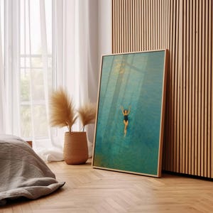May include: A framed art print depicts a person swimming in turquoise water. The swimmer is wearing a black swimsuit. The artwork is in a light wood frame and leans against a wooden wall. A woven basket with dried plants sits near a window.