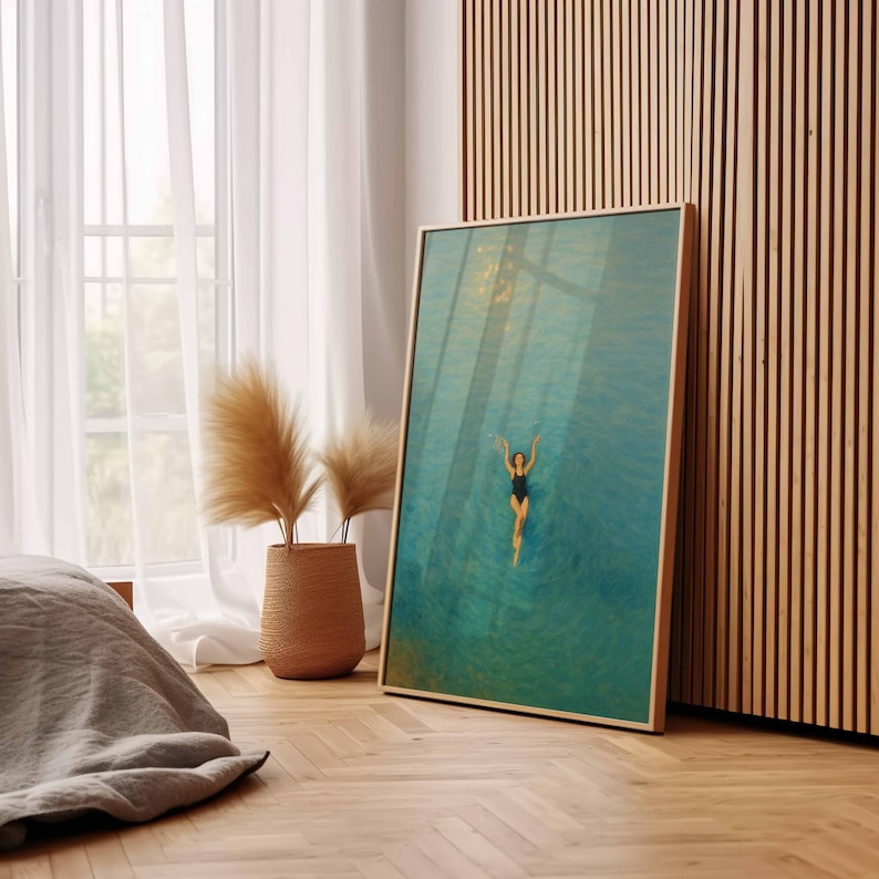 May include: A framed art print depicts a person swimming in turquoise water. The swimmer is wearing a black swimsuit. The artwork is in a light wood frame and leans against a wooden wall. A woven basket with dried plants sits near a window.