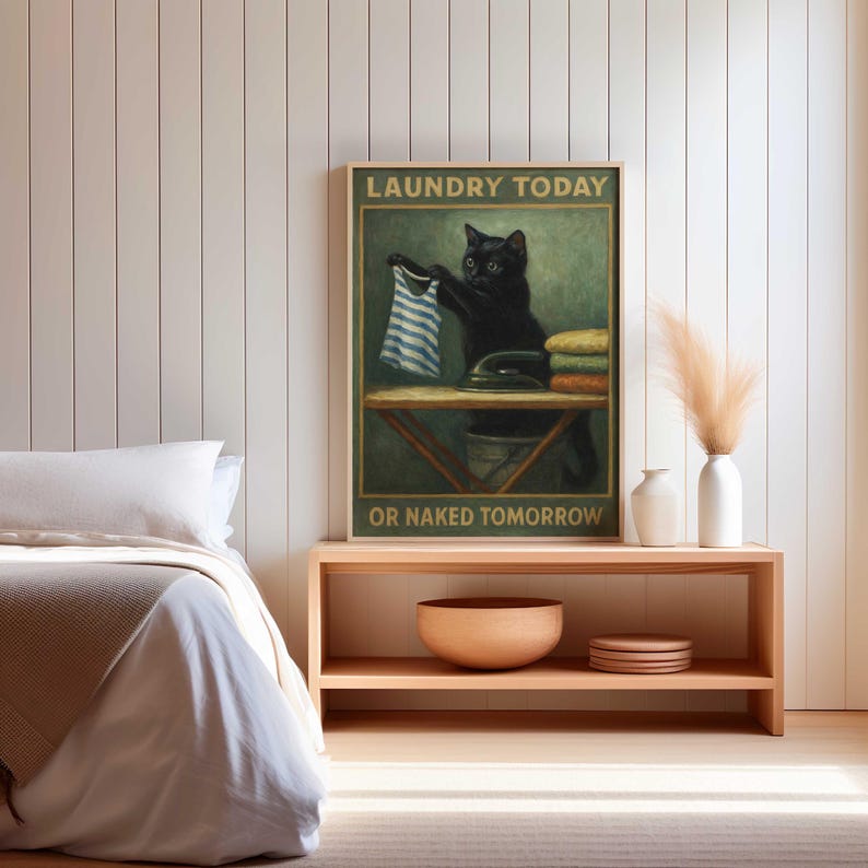 May include: A framed print featuring a black cat holding a striped shirt, with the text "LAUNDRY TODAY OR NAKED TOMORROW." The cat is depicted in a vintage style, with an ironing board and iron in the background. The print is displayed on a wooden shelf.