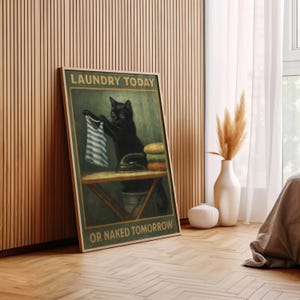 May include: Framed wall art featuring a black cat holding a blue and white striped shirt, with an iron and folded towels on a table. The text reads "LAUNDRY TODAY OR NAKED TOMORROW" in a vintage style.