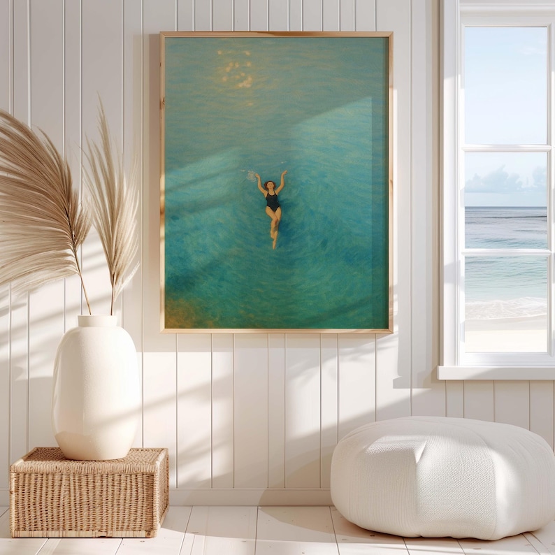May include: Framed art print featuring a person swimming in turquoise water. The artwork has a gold frame and is displayed on a white wall. A vase with dried plants and a woven basket are in the foreground.