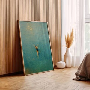 May include: A framed art print featuring a person swimming in turquoise water. The artwork has a light wooden frame and is leaning against a wooden wall. A white vase with dried plants and a white decorative object are nearby.