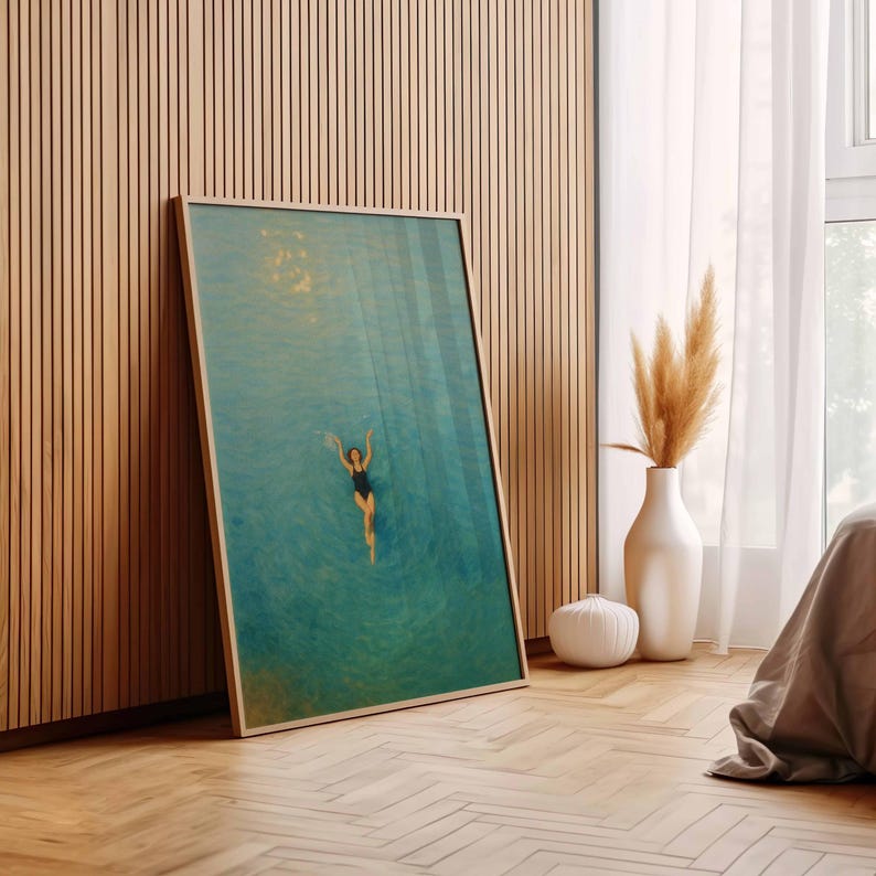 May include: A framed art print featuring a person swimming in turquoise water. The artwork has a light wooden frame and is leaning against a wooden wall. A white vase with dried plants and a white decorative object are nearby.